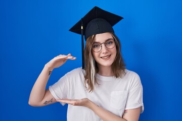 Blonde caucasian woman wearing graduation cap gesturing with hands showing big and large size sign, measure symbol. smiling looking at the camera. measuring concept.