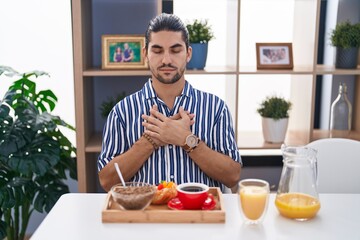 Hispanic man with long hair sitting on the table having breakfast smiling with hands on chest with closed eyes and grateful gesture on face. health concept.