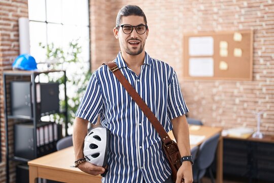 Hispanic man with long hair working at the office holding bike helmet winking looking at the camera with sexy expression, cheerful and happy face.