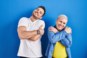 Young brazilian mother and son standing over blue background hugging oneself happy and positive,...