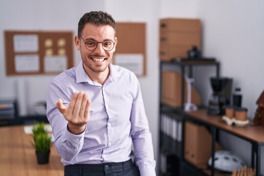 Young hispanic man at the office beckoning come here gesture with hand inviting welcoming happy and smiling