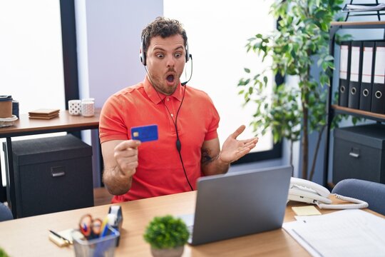 Young hispanic man wearing call center agent headset holding credit card scared and amazed with open mouth for surprise, disbelief face