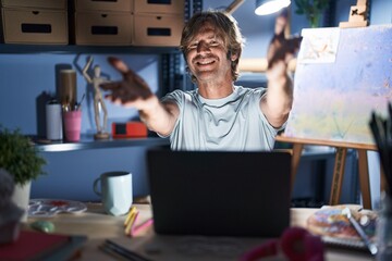 Middle age man sitting at art studio with laptop at night looking at the camera smiling with open arms for hug. cheerful expression embracing happiness.