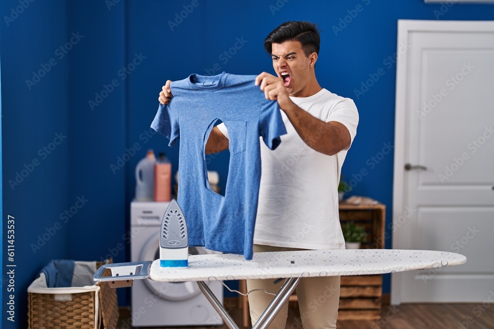 Canvas Prints Hispanic man ironing holding burned iron shirt at laundry room angry and mad screaming frustrated and furious, shouting with anger. rage and aggressive concept.