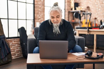 Middle age grey-haired man musician using laptop sitting on chair at music studio