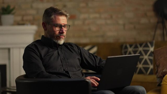 Mid adult businessman with laptop computer in home office, banking online, remote working. Portrait of older gray hair bearded man. Entrepreneur managing business on internet.