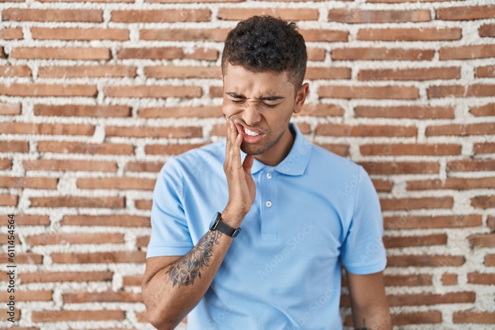 Poster brazilian young man standing over brick wall touching mouth with hand with painful expression becaus