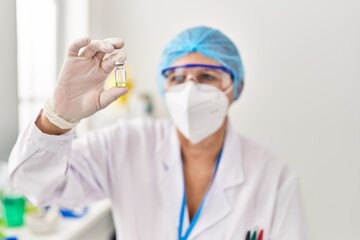 Naklejka premium Middle age woman wearing scientist unifor and medical mask holding dose vaccine at laboratory