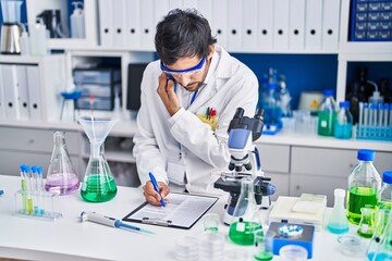 Young hispanic man scientist talking on the smartphone write on document at laboratory