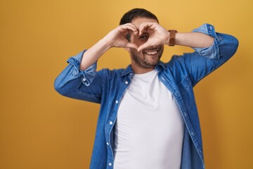 Hispanic man standing over yellow background doing heart shape with hand and fingers smiling...