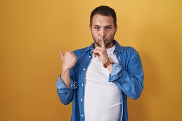 Hispanic man standing over yellow background asking to be quiet with finger on lips pointing with hand to the side. silence and secret concept.