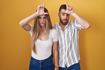 Young couple standing over yellow background making fun of people with fingers on forehead doing...