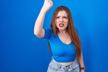Obraz na płótnie Canvas Redhead woman standing over blue background angry and mad raising fist frustrated and furious while shouting with anger. rage and aggressive concept.