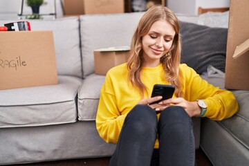 Young blonde woman using smartphone sitting on floor at new home