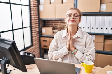 Young caucasian woman working at small business ecommerce using laptop praying with hands together asking for forgiveness smiling confident.