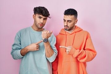 Young hispanic gay couple standing over pink background in hurry pointing to watch time,...