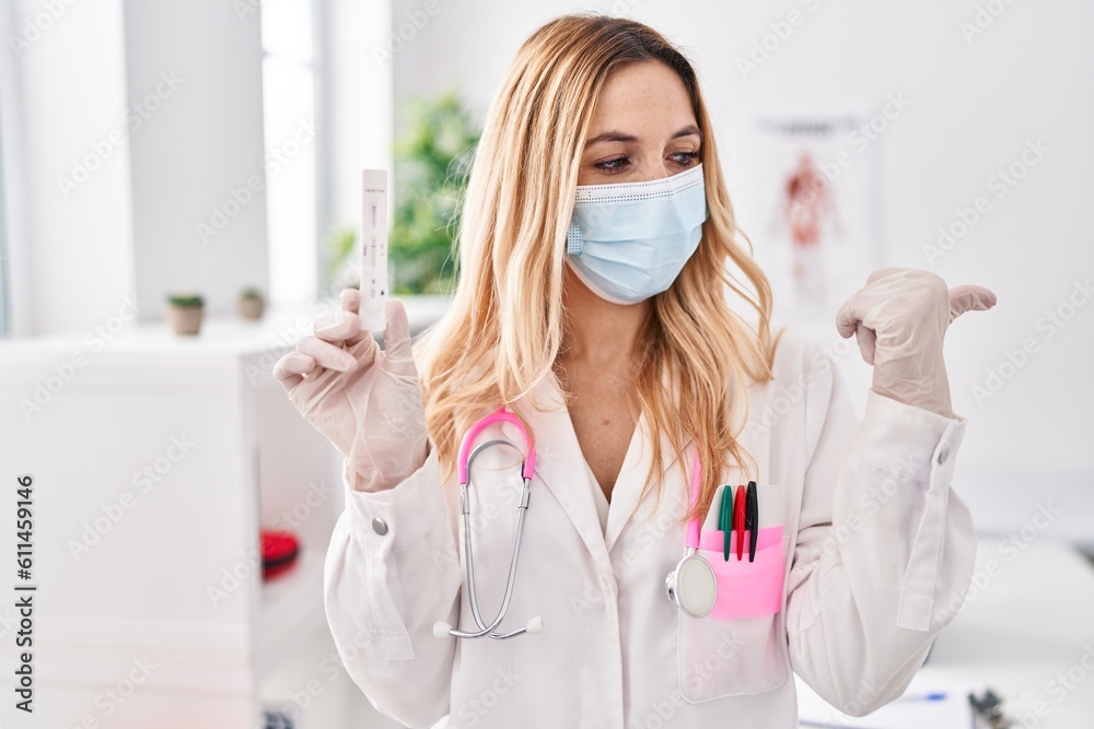 Sticker Young caucasian doctor woman holding covid test pointing thumb up to the side smiling happy with open mouth