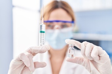 Young blonde woman scientist wearing medical mask holding covid-19 vaccine at laboratory