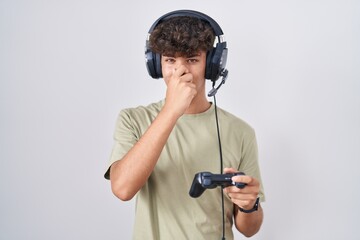 Hispanic teenager playing video game holding controller smelling something stinky and disgusting,...