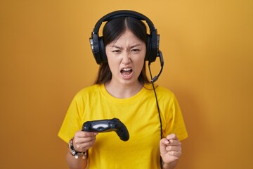 Chinese young woman playing video game holding controller angry and mad screaming frustrated and furious, shouting with anger. rage and aggressive concept.
