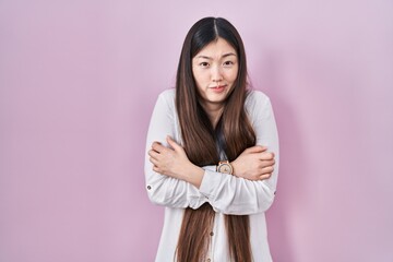 Chinese young woman standing over pink background shaking and freezing for winter cold with sad and...