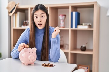 Obraz na płótnie Canvas Chinese young woman putting coin in piggy bank scared and amazed with open mouth for surprise, disbelief face