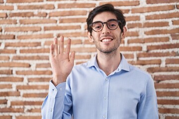 Young hispanic man standing over brick wall background waiving saying hello happy and smiling, friendly welcome gesture