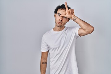 Hispanic man standing over isolated background making fun of people with fingers on forehead doing...
