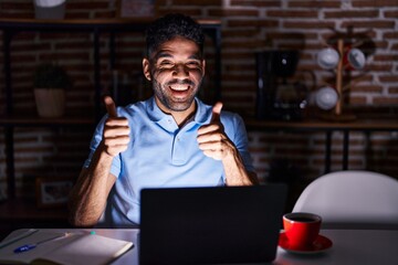 Fototapeta na wymiar Hispanic man with beard using laptop at night success sign doing positive gesture with hand, thumbs up smiling and happy. cheerful expression and winner gesture.