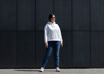 Full Length Pretty Mature Brunette Woman in White Hoodie and Sunglasses Looks Away, Dark Background Wall. Sportive Confident 40 Yo Beautiful Caucasian Woman in City. Horizontal Plane 