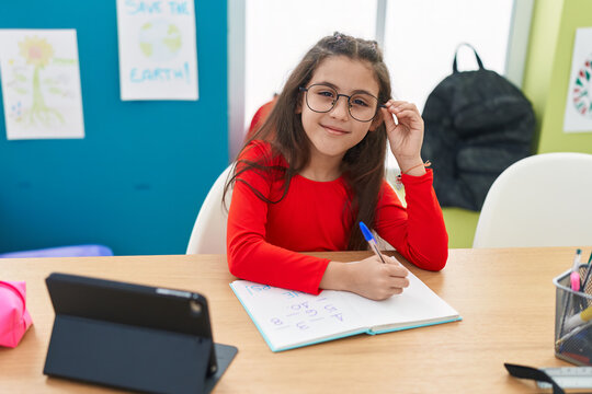Adorable hispanic girl student writing on notebook using touchpad at classroom