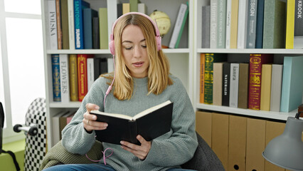 Young blonde woman student listening to music reading book at library university