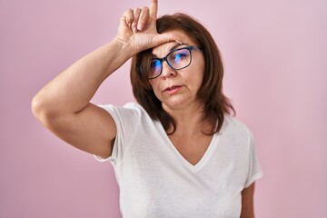 Middle age hispanic woman standing over pink background making fun of people with fingers on forehead doing loser gesture mocking and insulting.