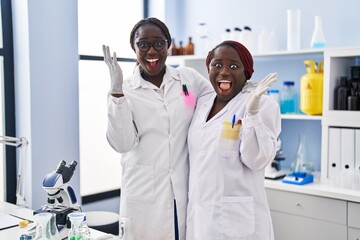 Two african women working at scientist laboratory celebrating victory with happy smile and winner expression with raised hands