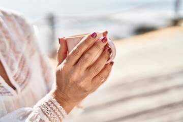 Middle age woman drinking coffee sitting on bench at seaside