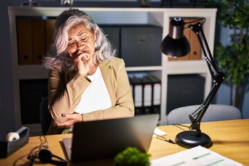 Middle age woman with grey hair working using computer laptop late at night thinking looking tired...