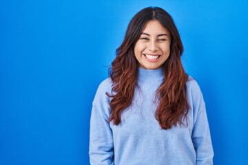 Hispanic young woman standing over blue background winking looking at the camera with sexy...