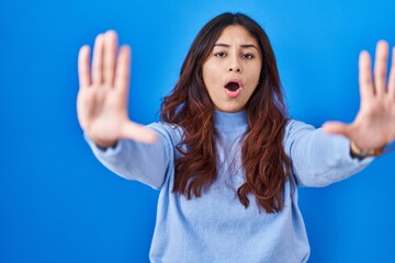 Fototapeta premium Hispanic young woman standing over blue background doing stop gesture with hands palms, angry and frustration expression