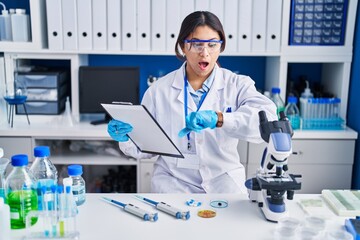 Hispanic young woman working at scientist laboratory looking at the watch time worried, afraid of getting late