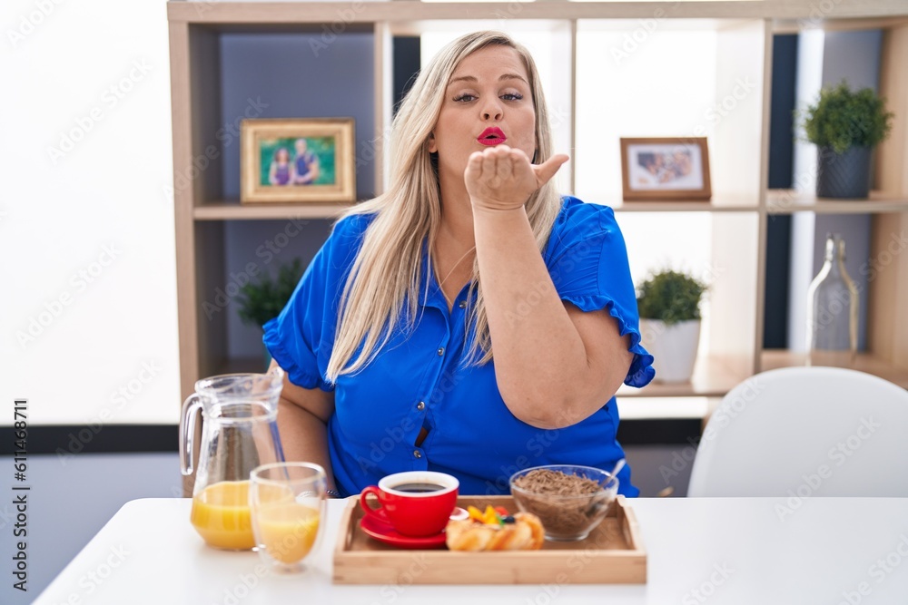 Poster caucasian plus size woman eating breakfast at home looking at the camera blowing a kiss with hand on