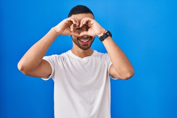 Naklejka premium Young hispanic man standing over blue background doing heart shape with hand and fingers smiling looking through sign