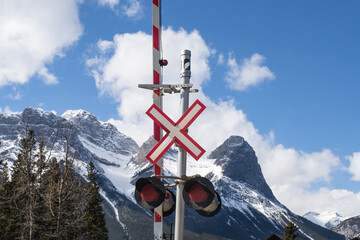 A railway level crossing gate and flashing lights with snow capped mountains and pine trees in the...