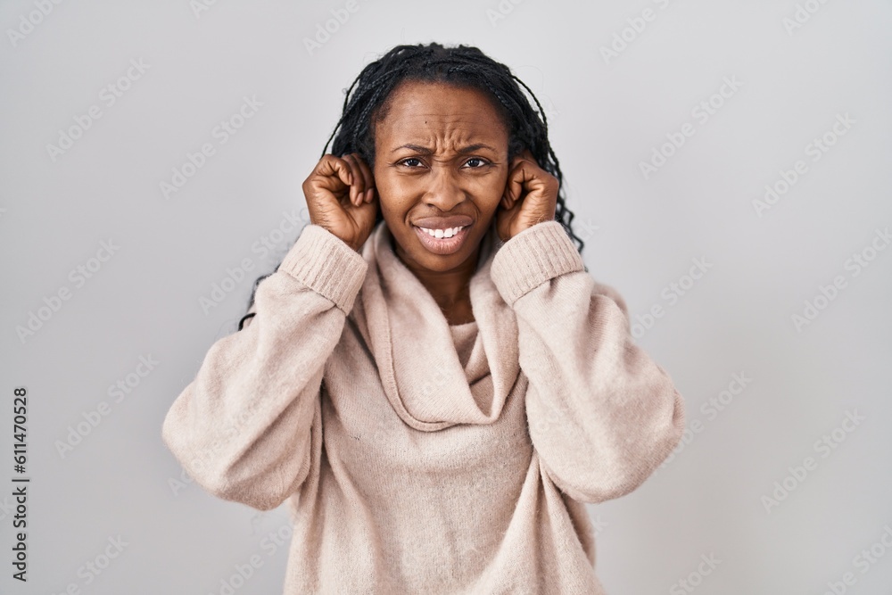 Sticker African woman standing over white background covering ears with fingers with annoyed expression for the noise of loud music. deaf concept.