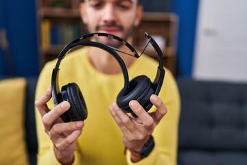 Young hispanic man holding broke headphones sitting on sofa at home