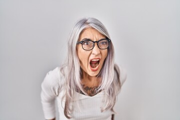 Middle age woman with grey hair standing over white background angry and mad screaming frustrated and furious, shouting with anger. rage and aggressive concept.