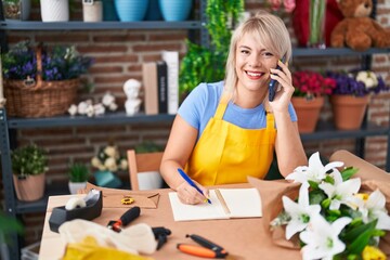 Young blonde woman florist talking on smartphone writing on notebook at florist