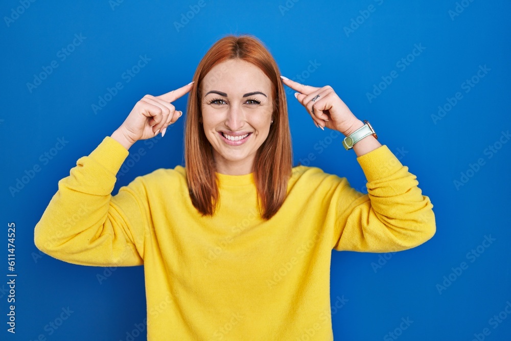 Poster Young woman standing over blue background smiling pointing to head with both hands finger, great idea or thought, good memory