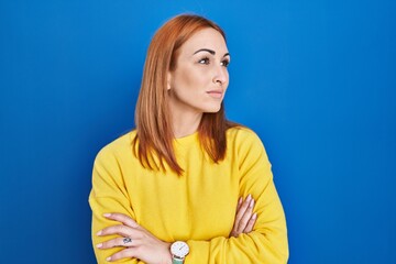 Young woman standing over blue background looking to the side with arms crossed convinced and confident