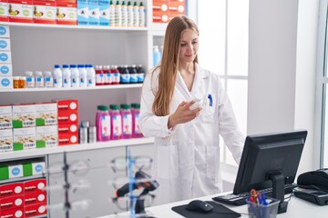 Young caucasian woman pharmacist holding pills bottle using computer at pharmacy