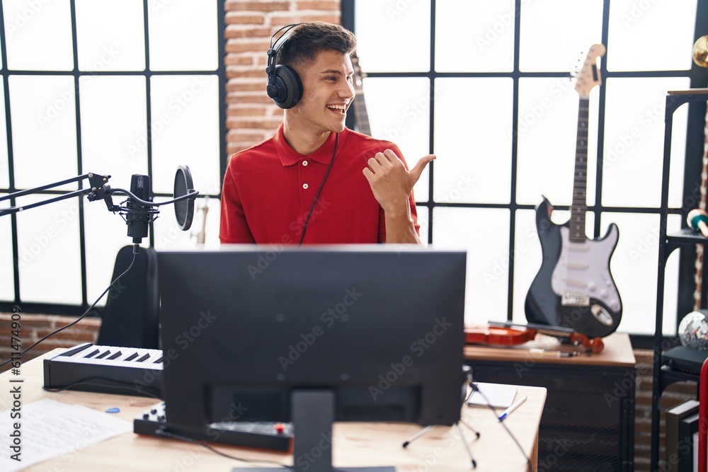 Wall mural Young hispanic man playing piano keyboard at music studio smiling with happy face looking and pointing to the side with thumb up.